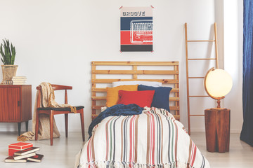 Striped sheets on wooden bed between ladder and chair in retro bedroom interior with poster. Real...