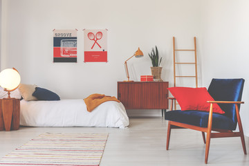 Red pillow on blue armchair in white bedroom interior with wooden cabinet next to bed. Real photo