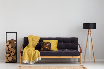 Log of wood next to comfortable sofa with yellow blanket and pillows, stylish wooden lamp with black lampshade, real photo copy space on the empty grey wall