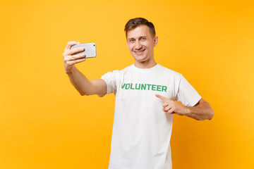 Portrait of man in white t-shirt written inscription green title volunteer taking selfie shot on mobile phone isolated on yellow background. Voluntary free assistance help charity grace work concept.