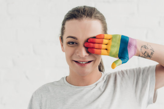 young transgender man covering eye with hand painted in colors of pride flag in front of white brick wall