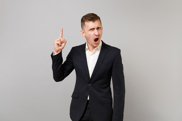 Outraged young business man in classic black suit, shirt swearing, pointing index finger up isolated on grey wall background in studio. Achievement career wealth business concept. Mock up copy space.