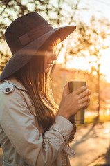 Young woman  in a hat with thermos thermo cup outdoor portrait in soft sunny daylight. Autumn. Sunset. Cozy