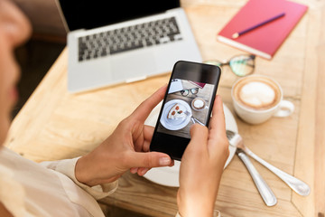 Cropped photo of european woman wearing hat photographing food on mobile phone, while sitting in stylish cafe