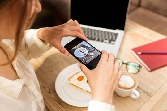 Cropped photo of young woman wearing hat photographing food on cell phone, while sitting in stylish cafe