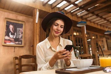Portrait of young woman wearing hat using smartphone, while sitting in stylish cafe