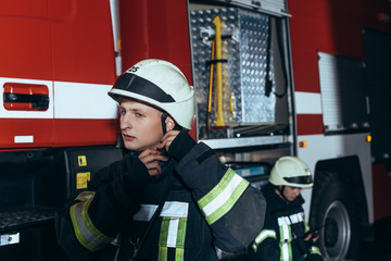 firefighter checking helmet with colleague behind at fire department