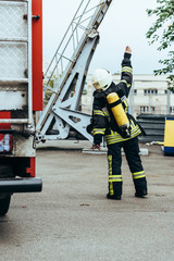 rear view of firefighter in uniform and helmet with fire extinguisher on back gesturing on street