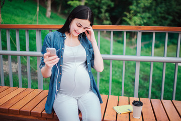 Fototapeta premium Portrait of a happy black hair and proud pregnant woman in a city in the background. She is sitting on a city bench. Expectant mother is listening to music in the park with an unborn child