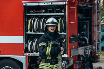portrait of fireman in protective uniform and helmet standing at truck with water hoses inside on street