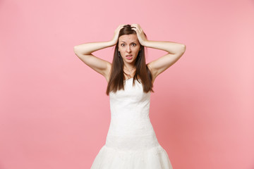 Portrait of bewildered puzzled bride woman in lace white wedding dress worries clinging to head standing isolated on pink pastel background. Wedding celebration concept. Copy space for advertisement.