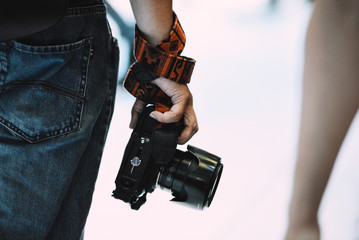 Close up standing unidentified man are holding the DSLR camera with one hand. The man wearing jeans looked from behind with the camera. Photographer hold camera real view