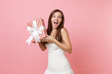 Portrait of surprised excited bride woman in lace white wedding dress holding red box with gift, present isolated on pink pastel background. Wedding celebration concept. Copy space for advertisement.