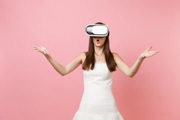 Portrait of shocked bride woman in white wedding dress, headset of virtual reality spreading hands isolated on pastel pink background. Organization of wedding concept. Copy space for advertisement.