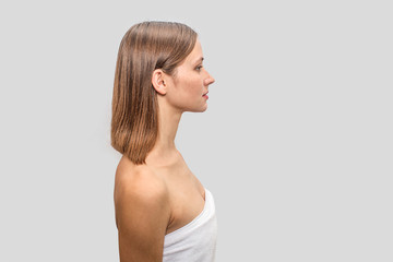 Picture of young woman stands side to camera and looks to right. She is serious. Body of young woman coveres with white towel. Isolated on grey background.