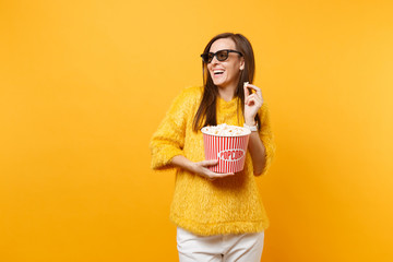 Laughing happy young girl in 3d imax glasses watching movie film, looking aside, holding bucket of popcorn isolated on bright yellow background. People sincere emotions in cinema, lifestyle concept.