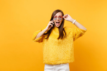 Amazed young woman holding heart glasses talking on mobile phone, conducting pleasant conversation isolated on bright yellow background. People sincere emotions, lifestyle concept. Advertising area.