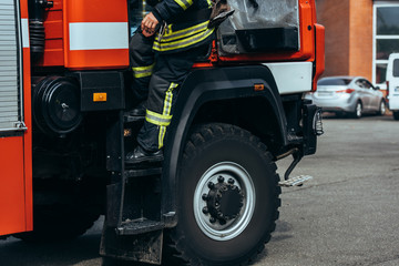 Obraz na płótnie Canvas cropped shot of firefighter in fireproof uniform standing on fire truck on street