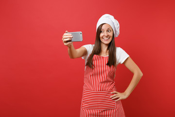 Housewife female chef cook or baker in striped apron, white t-shirt, toque chefs hat isolated on red wall background. Smiling fun woman doing selfie shot on mobile phone. Mock up copy space concept.