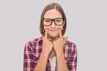 Bored but funny young woman puts fingers on her smiles. She looks exhausted. Model wears glasses. She looks smart. Isolated on grey background.