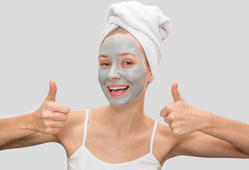 Positive and funny young woman shows her big thumbs up. Model smiles. She has mud mask on her face. Woman has white towel around her hair and t-shirt on body. Isolated on grey background.