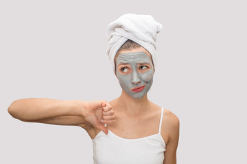 Unhappy and unsatisfied young woman looks up to left and shows her big thumb down. Model is upset. She has mud mask on her face, white towel around hair. Isolated on grey background.