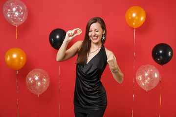 Joyful girl in black dress doing winner gesture holding bitcoin metal coin of golden color future currency on bright red background air balloon. Happy New Year, birthday mockup holiday party concept.