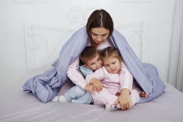 Fototapeta premium Young beautiful woman mother sitting under a blanket with children in bed with blue linens.