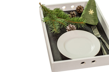 A round white plate, fork, knife, spruce branch, cones and a paper napkin folded in the shape of a Christmas tree on a rectangular wooden tray. 