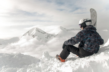 Fototapeta na wymiar Snowboarder holds snowboard in hands sits on big rock on mountains backdrop. Bansko, Bulgaria