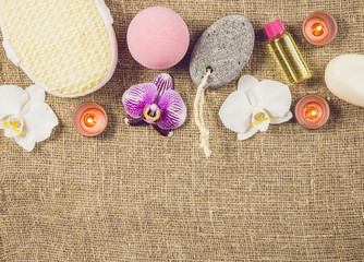 Fototapeta na wymiar Different spa products on rustic brown burlap sackcloth: pink bath bomb, white soap, massage oil, tea candles burning, gray pumice stone, sponge, blossoms, relaxing country home background concept.