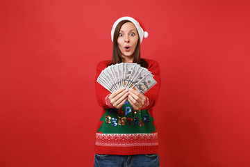 Amazed young Santa girl looking surprised, holding lots bunch of dollars banknotes, cash money isolated on red background. Happy New Year 2019 celebration holiday party concept. Mock up copy space.