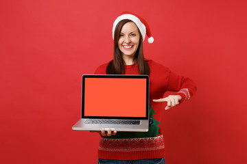 Cheerful young Santa girl pointing index finger on laptop pc computer with blank empty screen isolated on red background. Happy New Year 2019 celebration holiday party concept. Mock up copy space.