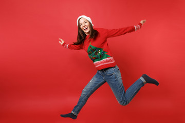 Laughing young Santa girl in knitted sweater, Christmas hat jumping, spreading hands, legs isolated on bright red background. Happy New Year 2019 celebration holiday party concept. Mock up copy space.