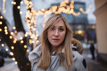 Winter portrait of happy young woman walking in snowy city decorated for Christmas and New Year holidays, wearing in a white coat with fur hood. Christmas lights on background. Black and white photo.