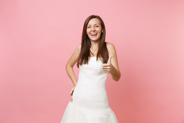 Portrait of laughing joyful bride woman in elegant white wedding dress standing and showing thumb up isolated on pink pastel background. Wedding celebration concept. Copy space for advertisement.