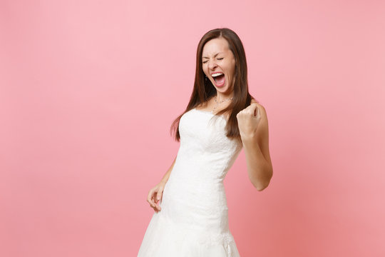 Portrait of overjoyed bride woman in white wedding dress standing doing winner gesture clenching fist isolated on pastel pink background. Wedding celebration concept. Copy space for advertisement.
