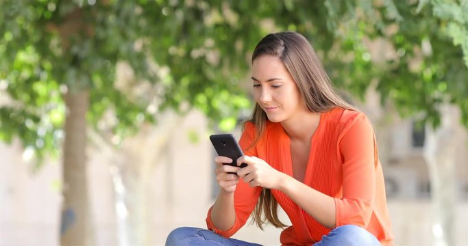Serious woman using a smart phone to send messages sitting on a bench in a park
