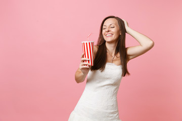 Portrait of smiling bride woman with closed eyes in white wedding dress keeping hand on head holding plastic cup with cola or soda isolated on pink pastel background. Wedding celebration. Copy space.