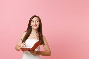 Dreamy bride woman in wedding dress looking up write note in notebook planning wedding choosing of staff wedding organizers isolated on pink pastel background. Wedding to do list. Celebration concept.
