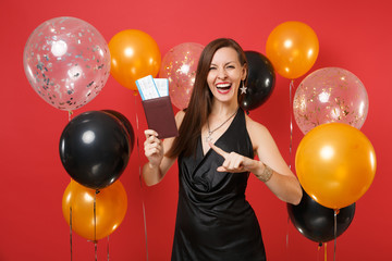 Laughing girl in black dress celebrating, pointing index finger on passport, boarding pass tickets in hand on bright red background air balloons. Happy New Year, birthday mockup holiday party concept.