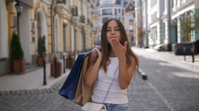 Lovely brunette girl carrying shopping bags over shoulders, turning back, blowing kiss and smling while walking along street after successful shopping. Portrait of cute young woman with shopping bags.