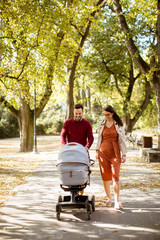 Happy young parents walking in the park and driving a baby in baby carriage
