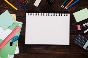 Empty spiral notebook with white page and multicolored stationery on brown wooden table. Workplace with notepad on colored background. Sketch-book for input the text, top view. Back to school concept