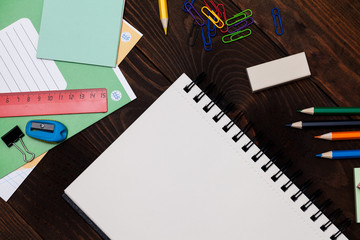 Empty spiral notebook with white page and multicolored stationery on brown wooden table. Workplace with notepad on colored background. Sketch-book for input the text, top view. Back to school concept