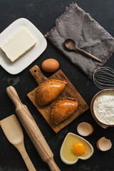 Fresh baked pasties and baking ingredients on a dark stone background. Russian pirozhki. Top view