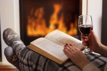 Young woman reading book and drinking red wine in front of the fireplace and warming feet in wool socks. Girl legs is covered with blanket. Winter long cold night concept. Close up, selective focus