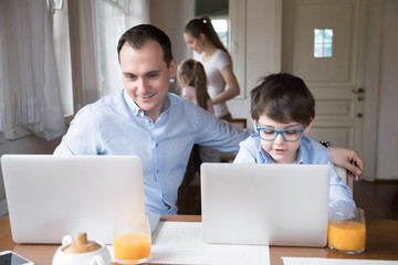 Happy family with small children spend free time on weekend together at home. Little son copying father sitting at the table using computers reading news having fun, mother and daughter on background