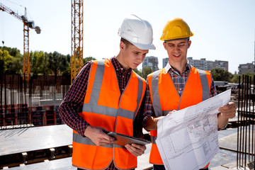 Structural engineer and construction manager dressed in orange work vests and helmets discuss documentation on the open air building site next to the crane