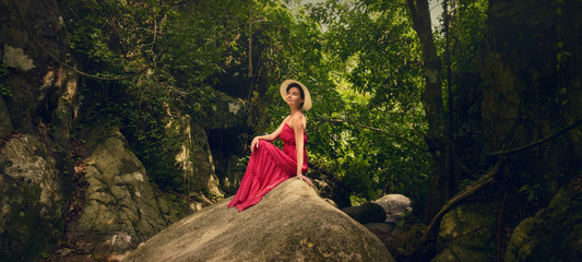 A girl in a hat sits on a rock in the jungle. Girl in a red dress. Thailand.